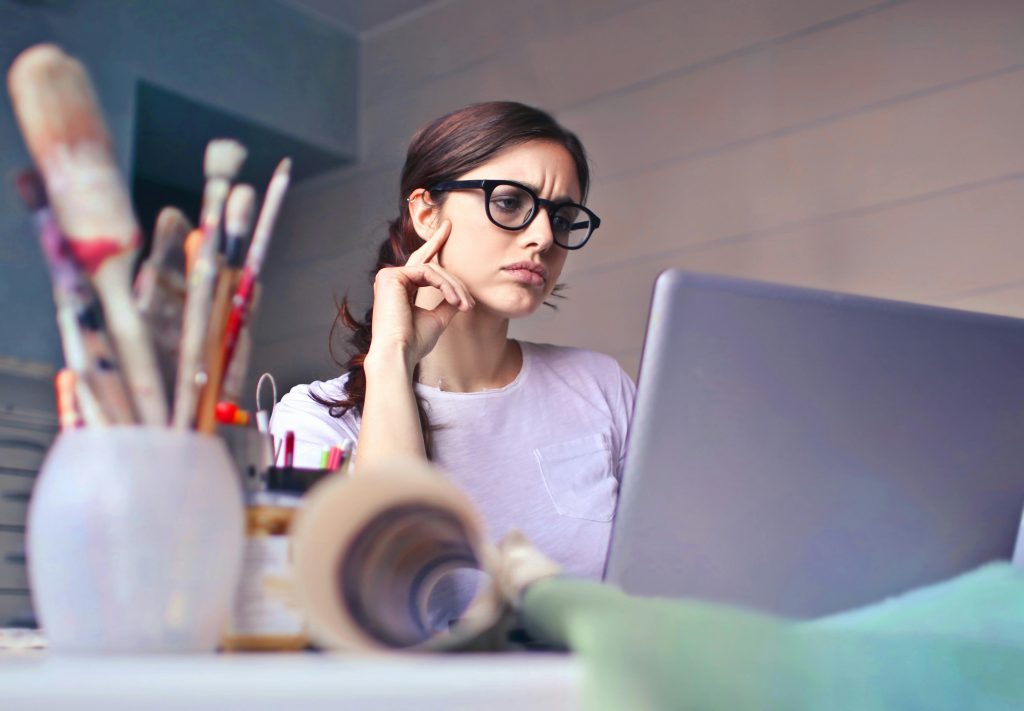 Focused woman with glasses working on laptop in an artistic workspace with paintbrushes.
